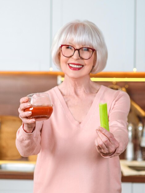 mulher sênior alegre com suco de aipo e tomate fazendo dieta na cozinha