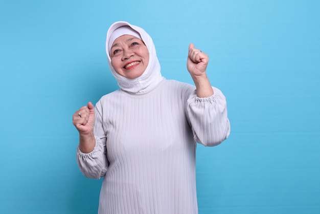 Mulher sênior animada em hijab branco sorrindo e comemorando a vitória com os punhos cerrados e levantada