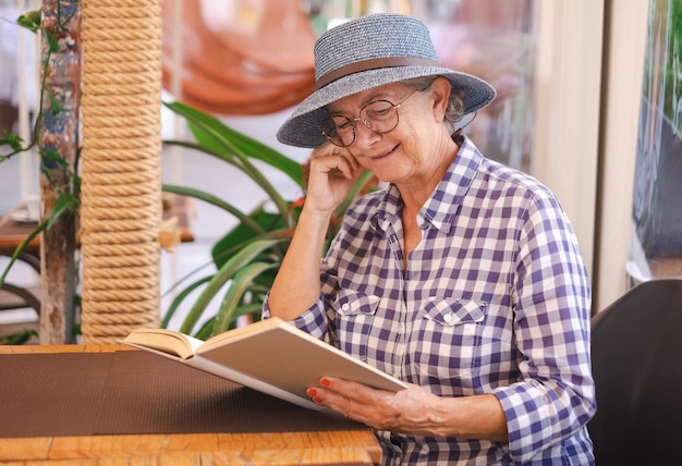 Mulher sênior atraente com chapéu sentado na mesa de café lendo um livro senhora caucasiana aproveitando o tempo livre e aposentadoria