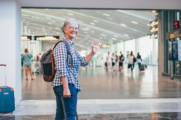 Mulher sênior atraente e feliz caminhando na área do aeroporto em direção ao portão de embarque acenando com a mão