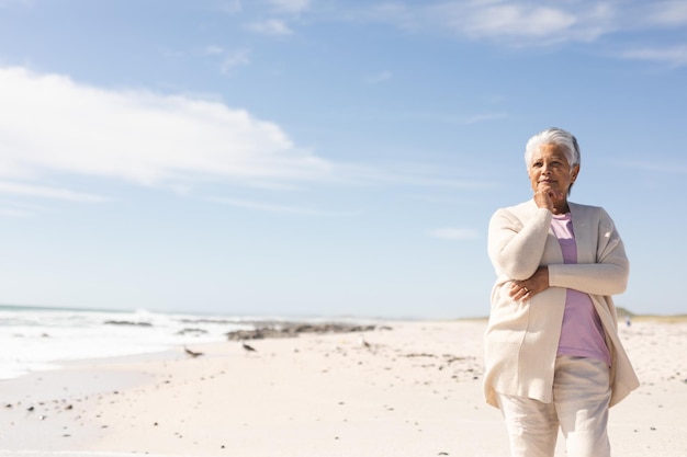 Mulher sênior birracial aposentada com a mão no queixo contemplando na praia contra o céu durante o dia ensolarado
