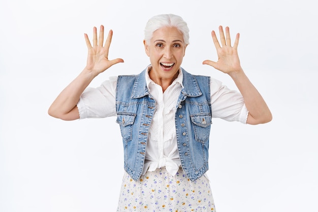 Mulher sênior bonita, feliz, entusiasmada, com cabelos grisalhos, usar colete jeans estiloso, vestido, levantando as mãos, mostrando o número dez, mais cinco, sorrindo alegremente