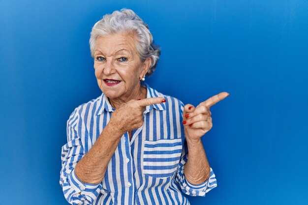 Foto mulher sênior com cabelos grisalhos em pé sobre fundo azul sorrindo e olhando para a câmera apontando com as duas mãos e os dedos para o lado.