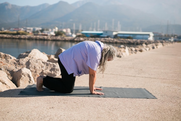 Mulher sênior com dreadlocks em posição de alongamento à beira-mar de manhã Mulher idosa fazendo ioga perto da praia