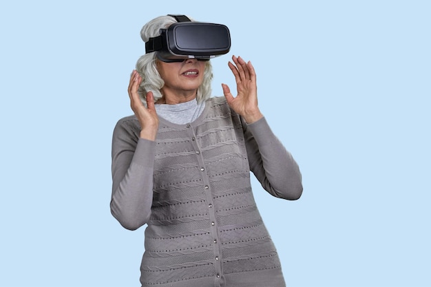 Mulher sênior com fone de ouvido de realidade virtual em fundo azul velha atordoada pela experiência vr