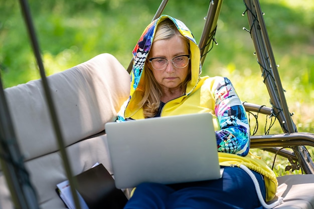 Mulher sênior com laptop e documentos trabalhando no jardim no conceito de escritório em casa verde do sofá de balanço
