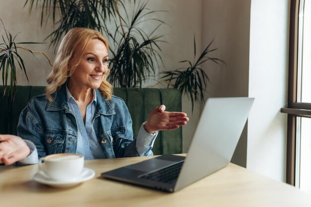 Mulher sênior de 50 anos está trabalhando on-line no caffe Ela está sentada na frente de um monitor de laptop e falando em videochamada on-line Conceito Freelance