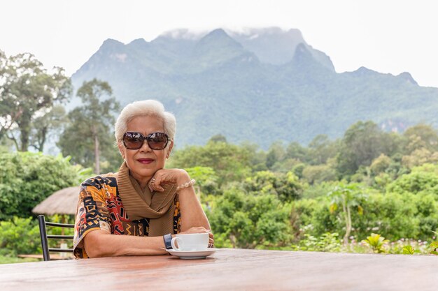Mulher sênior de cabelos brancos grisalhos tomando café no café ao ar livre com vista para a montanha
