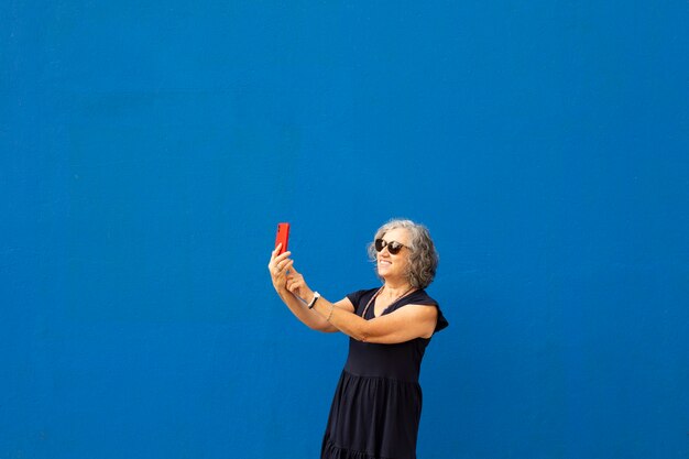 Mulher sênior de cabelos grisalhos tirando uma selfie com um smartphone vermelho contra uma parede azul