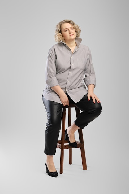 Mulher sênior de camisa cinza e calças de couro falso senta-se na cadeira de madeira alta sobre fundo cinza do estúdio