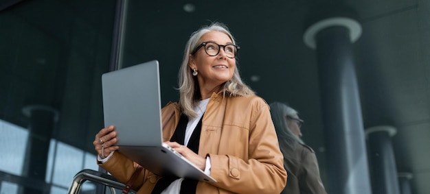 Foto mulher sênior de negócios trabalhando em um laptop online contra o pano de fundo de um prédio de escritórios