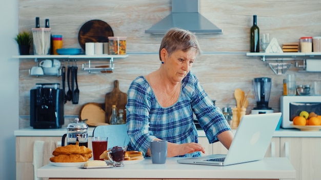 Mulher sênior em busca de receita on-line usando o laptop na cozinha durante o café da manhã. Idoso aposentado trabalhando em casa, teletrabalho usando comunicação on-line de trabalho remoto da Internet em tecnologia moderna