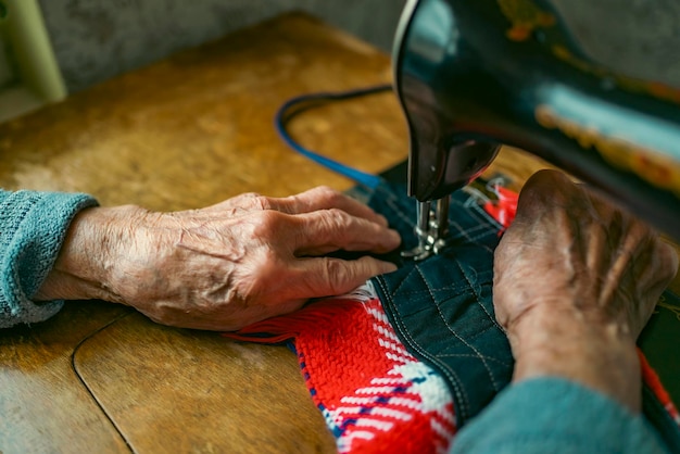Mulher sênior em óculos usa máquina de costura mãos enrugadas da velha costureira mulher idosa Máquina de costura antiga Máquina de costura manual clássica estilo retrô pronta para o trabalho de costura