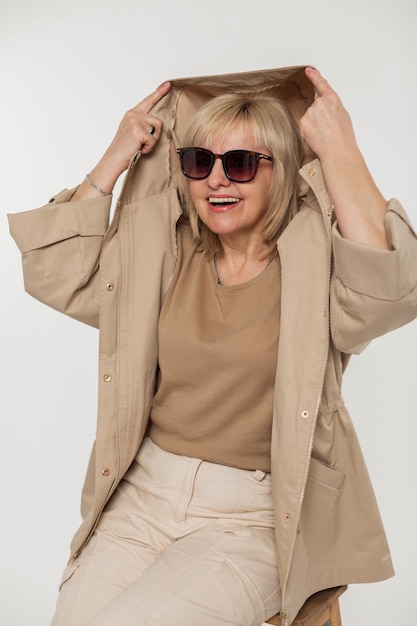 Mulher sênior engraçada e feliz com óculos de sol vintage legais em roupa bege da moda com uma jaqueta coloca um capuz e se diverte em um fundo branco