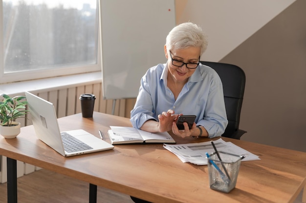 Mulher sênior europeia elegante e confiante usando smartphone no local de trabalho Empresária elegante e madura de cabelos grisalhos dos anos 60 com telefone celular no escritório Líder chefe usando aplicativos de internet