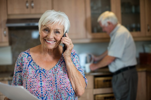 Mulher sênior falando no celular enquanto um homem trabalha na cozinha de casa