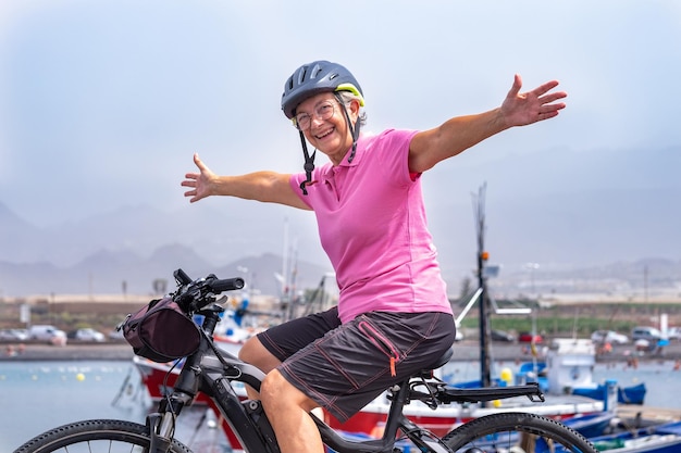 Mulher sênior feliz ativa com capacete correndo sua bicicleta elétrica ao longo do mar com os braços abertos