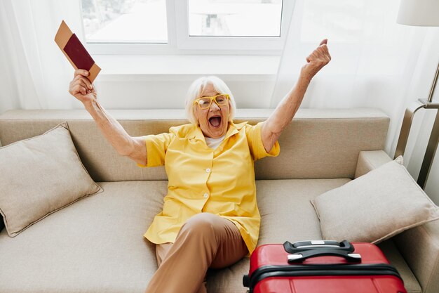 Mulher sênior feliz com passaporte e bilhete de viagem embalado uma mala vermelha férias e cuidados de saúde sorridente velha sentada alegremente no sofá antes da viagem levantou as mãos de alegria