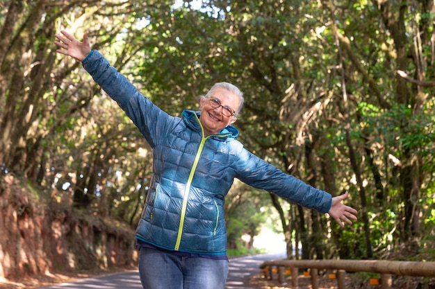 Foto mulher sênior feliz nas montanhas sob a sombra das árvores olhando para a câmera com os braços abertos