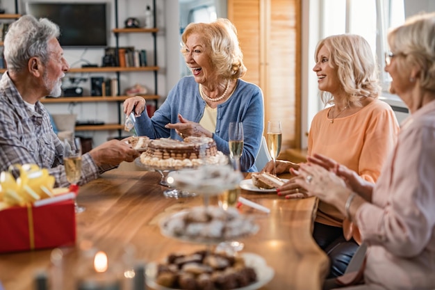 Mulher sênior feliz se divertindo em sua festa de aniversário enquanto serve um bolo e se comunica com amigos em casa