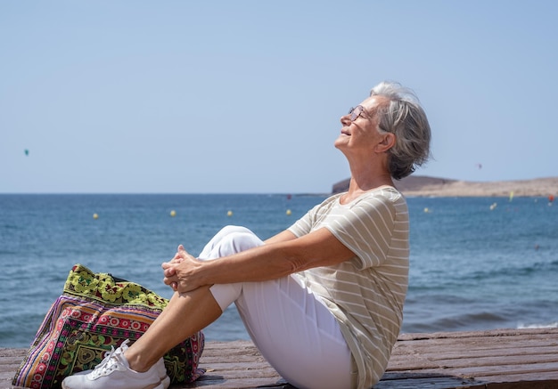 Mulher sênior feliz sentada perto da praia com o rosto virado para o sol senhora bonita idosa relaxa enquanto aproveita as férias na praia em um dia ensolarado e ventoso