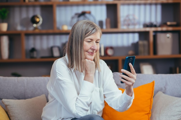 Mulher sênior feliz usa telefone para comunicação online sentada no sofá em casa à noite e sorrindo