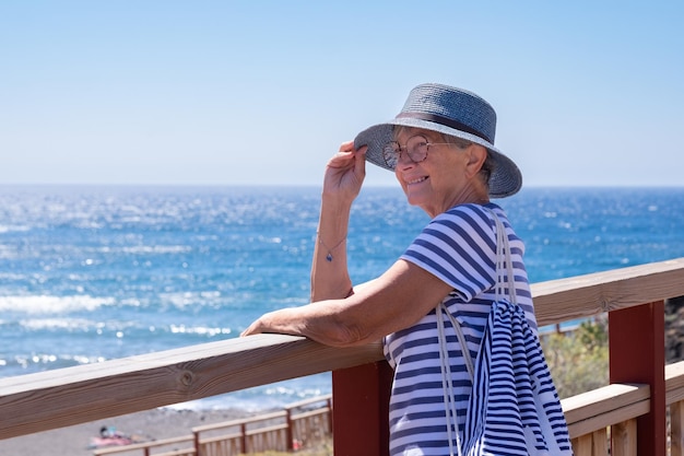 Mulher sênior feliz vestida de azul andando ao longo do mar segurando seu chapéu para que não voe Senhora madura com óculos desfrutando de liberdade e relaxe nas férias