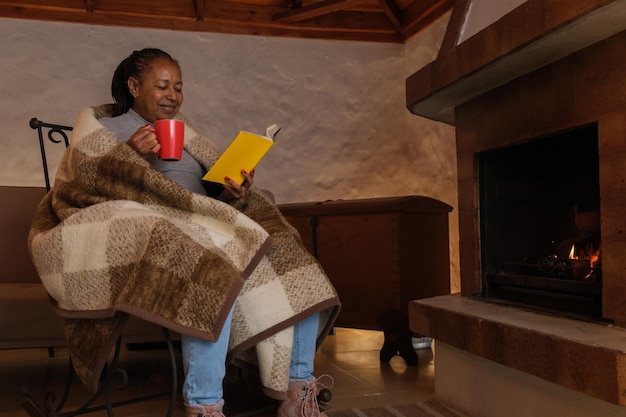 Mulher sênior lendo um livro em uma cadeira de balanço ao lado da lareira tendo um conceito de chocolate quente relaxamento inverno aconchegante