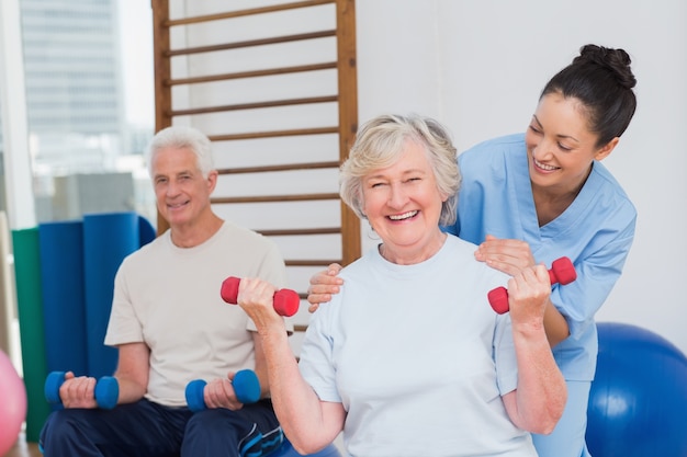 Mulher sênior levantando dumbbells enquanto está sentado com homem e treinador