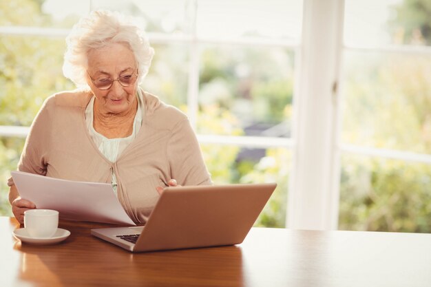 Mulher sênior, lidar com documentos enquanto estiver usando o laptop em casa