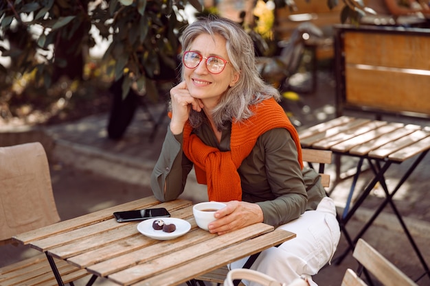 Mulher sênior positiva com óculos e uma xícara de café na mesa no terraço do café ao ar livre