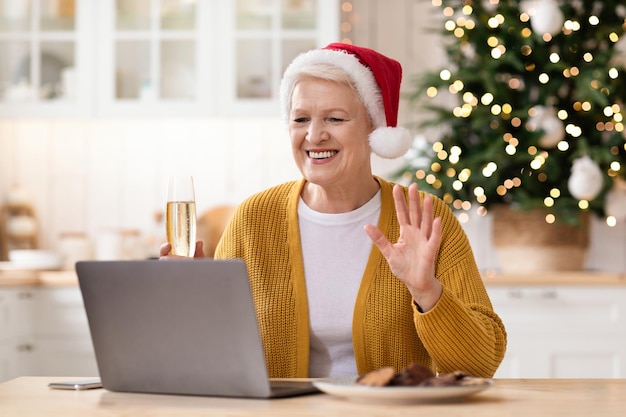 Mulher sênior positiva no chapéu de Papai Noel, bebendo champanhe e tendo vídeo-conferência com a família dela, sentada na cozinha decorada e usando o laptop, acenando para a câmera. Comemorando o natal ou ano novo