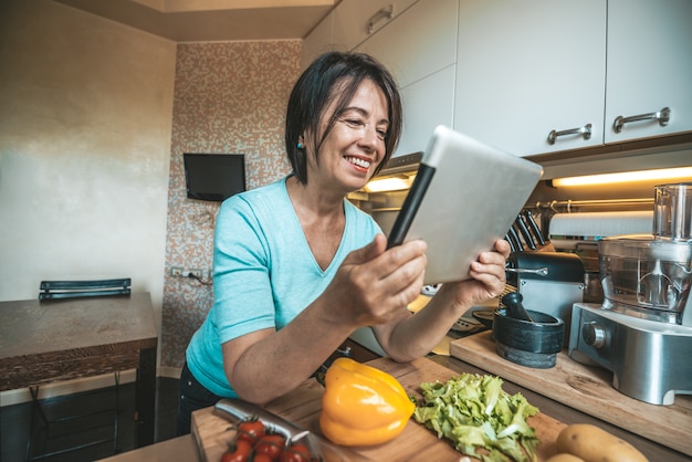 Foto mulher sênior que cozinha vegetais em casa lendo uma receita sobre o tablet.