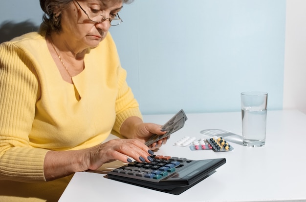 Mulher sênior segurando dinheiro e contando com a calculadora. pensionista sentado na cozinha com medicamentos. conceito de depressão, preço do medicamento, custo do tratamento