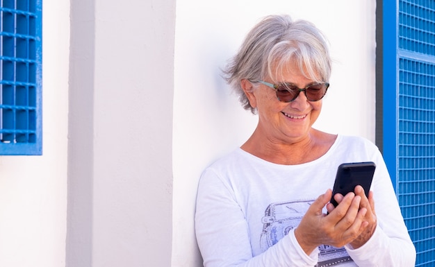 Mulher sênior sorri olhando seu smartphone. em pé contra uma parede branca usando tecnologia sem fio. estilo de vida alegre para idosos