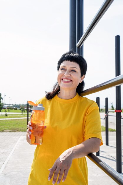 Mulher sênior sorridente bebendo água após treino ao ar livre no campo de esportes