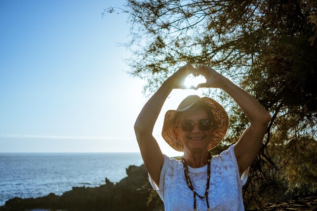 Foto mulher sênior sorridente com chapéu de palha de pé perto do mar ao pôr do sol fazendo forma de coração com as mãos