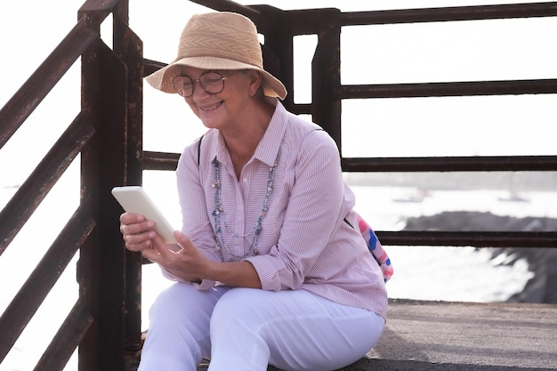 Mulher sênior sorridente com chapéu sentado ao ar livre no mar usando celular