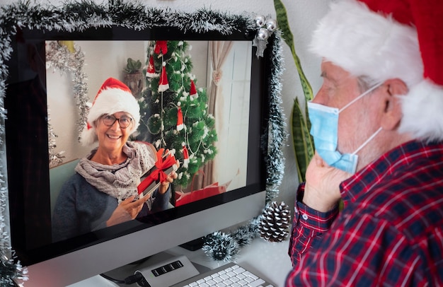 Mulher sênior sorridente falando na chamada de vídeo para cumprimentos de Natal, mostrando o presente recebido. Homem com chapéu de Papai Noel usa máscara cirúrgica devido à quarentena por infecção por coronavírus