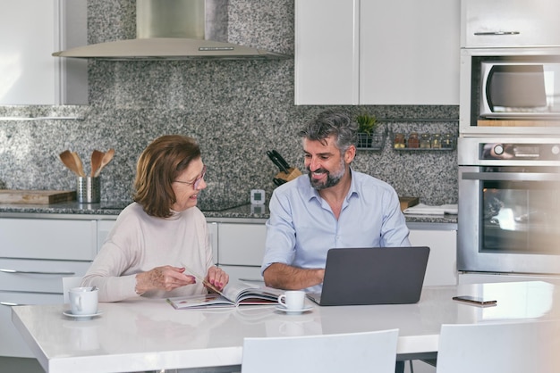 Mulher sênior sorridente lendo uma revista e olhando para um homem maduro digitando no laptop enquanto está sentada na mesa da cozinha em casa