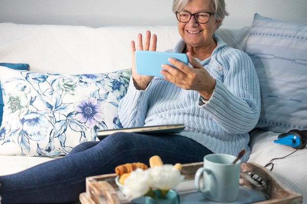 Mulher sênior sorridente relaxando no sofá em casa segurando smartphone em videochamada com família ou amigos