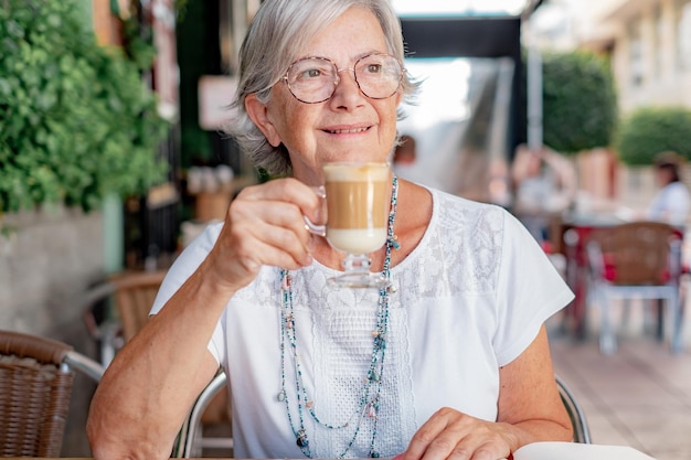 Mulher sênior sorridente sentada ao ar livre em uma mesa de café desfrutando de uma bebida de café e leite senhora idosa caucasiana com óculos relaxada na aposentadoria ou férias