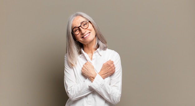 Mulher sênior sorrindo alegremente e comemorando, com os punhos cerrados e os braços cruzados, sentindo-se feliz e positiva