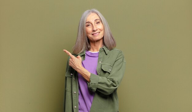 Mulher sênior sorrindo alegremente, sentindo-se feliz e apontando para o lado e para cima, mostrando o objeto no espaço da cópia