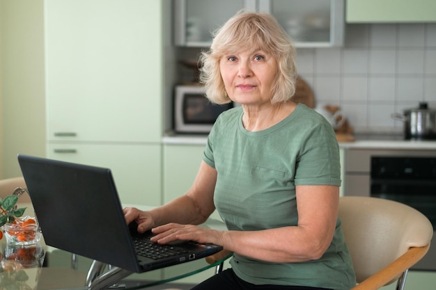 Mulher sênior trabalhando no laptop sentado na cozinha olhando para a câmera