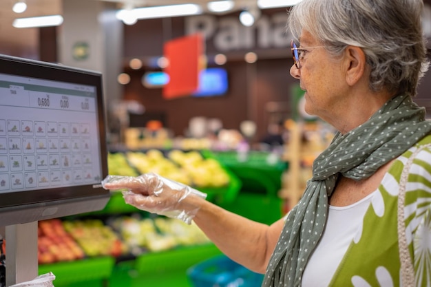 Mulher sênior usando luvas de proteção pesa suas compras de legumes na balança eletrônica do supermercado