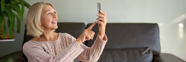 Mulher sênior usando telefone celular enquanto está sentado no sofá. Mulher mais velha sentada no sofá e mandando mensagem de texto uma mensagem telefônica.
