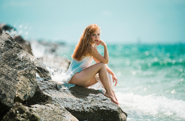 Mulher sensual sentada na praia rochosa de areia contra as ondas azuis menina de verão sonho jovem atraente