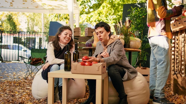 Mulher sentada à mesa com enólogo degustando vinho orgânico caseiro, apreciando a amostragem do produto antes de comprá-lo. Compras no mercado de agricultores do festival de comida de rua natural.