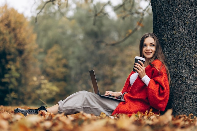 Mulher sentada debaixo da árvore no parque trabalhando no laptop e tomando café